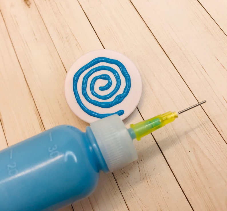 Op de afbeelding: Een witte keramische schijf met een blauw spiraalontwerp erop geschilderd. Een blauwe plastic fles met een gele punt wordt gebruikt om de verf aan te brengen.