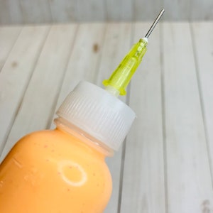 May include: A clear plastic bottle with a white cap and a yellow needle attached. The bottle is filled with a light orange liquid.