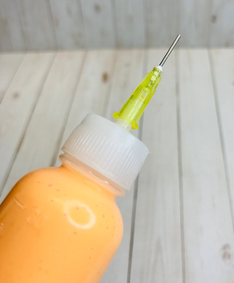 May include: A clear plastic bottle with a white cap and a yellow needle attached. The bottle is filled with a light orange liquid.