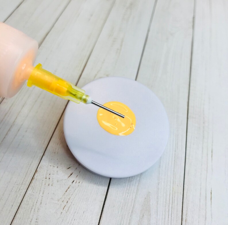 May include: A close-up of a syringe dispensing yellow paint onto a white circular surface. The syringe has a clear barrel and a yellow plunger. The paint is forming a small circle in the center of the surface.