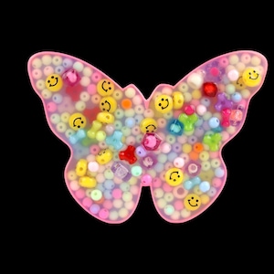 Può includere: Un oggetto in resina rosa a forma di farfalla riempito di perline colorate e perline a forma di faccina sorridente. Le perline sono di vari colori, tra cui giallo, rosa, blu e rosso. Lo sfondo è nero.