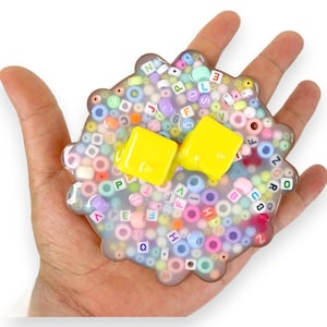 May include: A clear slime with colorful alphabet beads and two yellow square-shaped objects inside. The slime is in a hand.