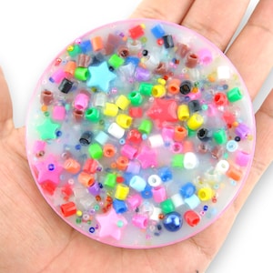 Puede incluir: Un posavasos redondo de resina transparente con borde rosa, lleno de cuentas de colores, estrellas y purpurina.