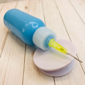 Op de afbeelding: Een blauwe plastic fles met een gele naald die aan de bovenkant is bevestigd. De naald rust op een witte plastic schijf.