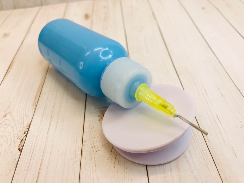 Op de afbeelding: Een blauwe plastic fles met een gele naald die aan de bovenkant is bevestigd. De naald rust op een witte plastic schijf.