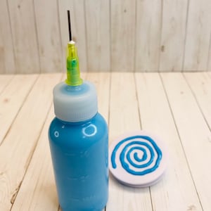 May include: A blue bottle with a needle tip and a white cap. The bottle is filled with blue paint. A white circle with a blue spiral design is next to the bottle.