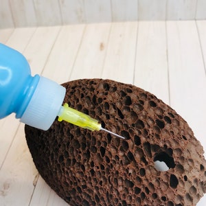 Op de afbeelding: Een blauwe plastic fles met een gele naald die in een bruine poreuze steen is gestoken.