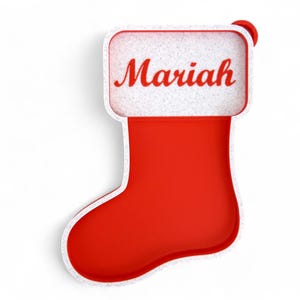 Peut inclure: Une chaussette de Noël rouge avec le nom "Mariah" en écriture rouge. La partie supérieure est blanche avec un aspect texturé. La chaussette a une bordure blanche et une petite boucle rouge.