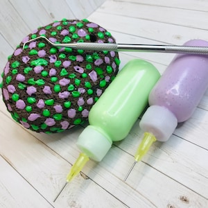 Puede incluir: Dos botellas de pintura con aplicadores de punta fina, una verde y otra morada, y una herramienta de metal para hacer puntos de pintura en una roca con puntos verdes y morados.