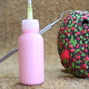 以下が含まれることがあります： 茶色の表面に、針状のアプリケーターが付いたピンクのボトル、金属製のツール、赤と緑の斑点のある丸い物体がある。