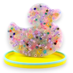 Op de afbeelding: Een helder paarse eend van hars met een gele basis. De eend is gevuld met kleurrijke kralen, glitter en andere kleine decoratieve items.