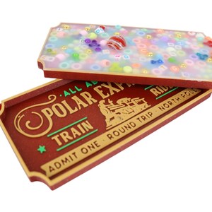Può includere: Un biglietto rosso e dorato con il testo "All Aboard Polar Express Train" e "Admit One Round Trip North Pole Ride". Il biglietto è aperto per rivelare una scatola trasparente piena di perline colorate.