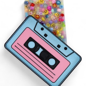 Puede incluir: Una bandeja con forma de cinta de cassette retro en azul claro y rosa, con detalles negros. Al lado, una bandeja rectangular llena de cuentas de colores, caras sonrientes y letras. Los objetos están sobre un fondo blanco.