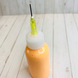 May include: A small, clear plastic bottle with a white cap and a yellow needle attached. The bottle is filled with a light orange liquid.