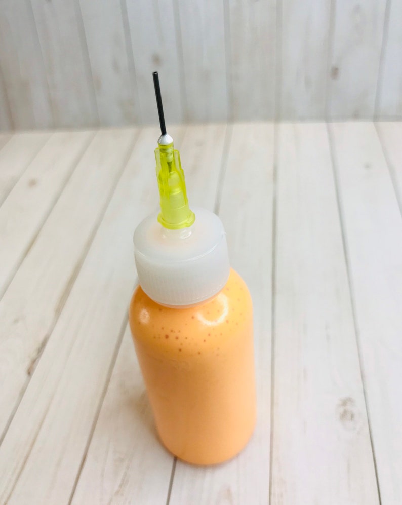 May include: A small, clear plastic bottle with a white cap and a yellow needle attached. The bottle is filled with a light orange liquid.