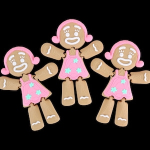 Puede incluir: Tres figuras de niña de pan de jengibre con gorros rosas y vestidos adornados con estrellas azules. Las niñas de pan de jengibre tienen detalles de glaseado blanco para sus caras, brazos y piernas. El fondo es negro liso.