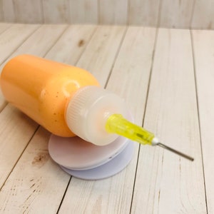May include: A small orange bottle with a clear cap and a yellow needle tip. The bottle is sitting on a white surface.