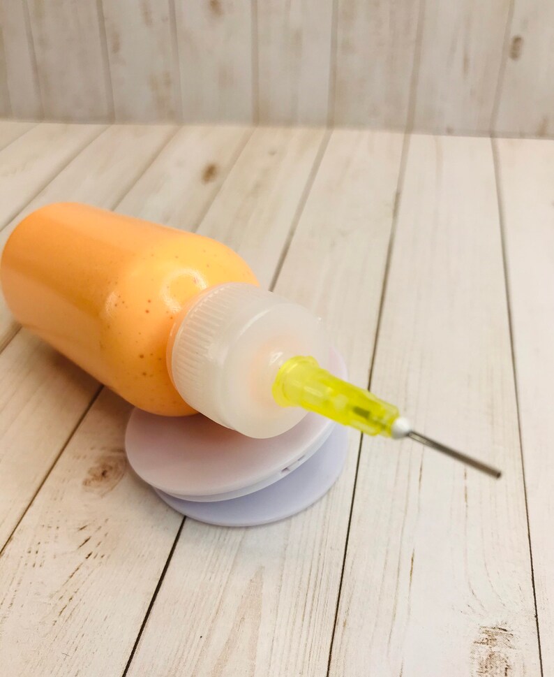 May include: A small orange bottle with a clear cap and a yellow needle tip. The bottle is sitting on a white surface.