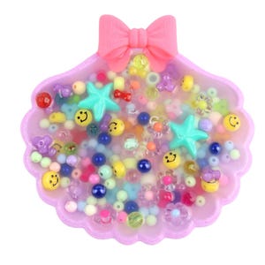 Pode incluir: Um recipiente rosa em forma de concha cheio de contas e berloques coloridos. O recipiente tem um laço rosa no topo. As contas incluem rostos sorridentes, estrelas e formas de flores. As contas são de várias cores, incluindo azul, amarelo e verde.