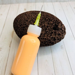 May include: A clear plastic bottle with a yellow needle attached to the top. The bottle is filled with a light orange liquid. The bottle is sitting on top of a brown pumice stone.