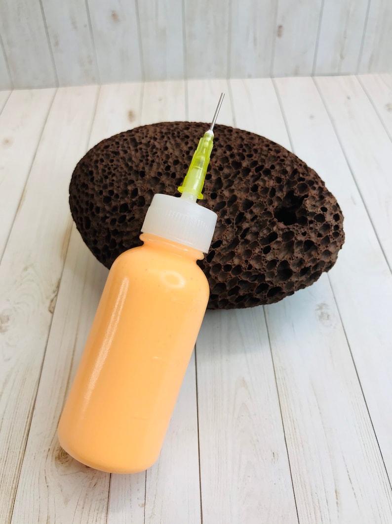 May include: A clear plastic bottle with a yellow needle attached to the top. The bottle is filled with a light orange liquid. The bottle is sitting on top of a brown pumice stone.