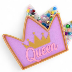 Può includere: Una decorazione a forma di corona color lavanda con un bordo dorato e un disegno a corona dorata. La parola "Queen" è scritta in rosa. La corona è riempita di perline colorate e piccoli abbellimenti.