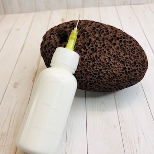 Op de afbeelding: Een witte plastic fles met een gele naald die aan de bovenkant is bevestigd. De fles staat op een lichtbruin houten oppervlak naast een bruine puimsteen.