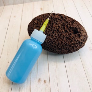 Op de afbeelding: Een lichtblauwe fles met een naald bevestigd aan een bruine puimsteen. De fles heeft een witte dop en een doorzichtige plastic buis.