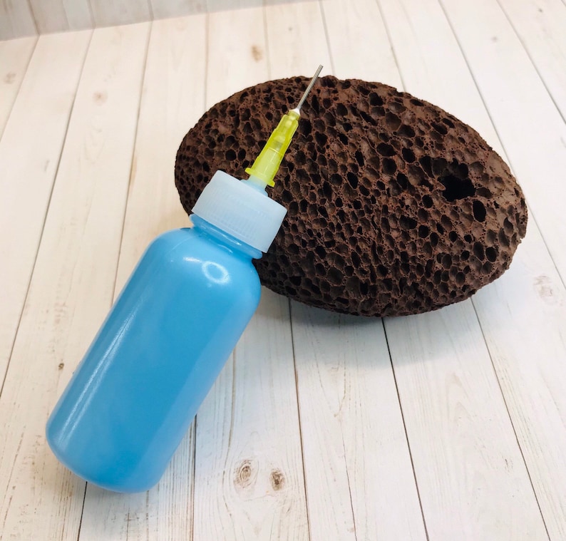 Op de afbeelding: Een lichtblauwe fles met een naald bevestigd aan een bruine puimsteen. De fles heeft een witte dop en een doorzichtige plastic buis.
