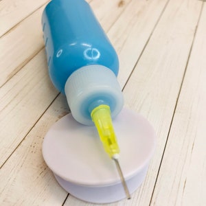 Op de afbeelding: Een blauwe plastic fles met een witte dop en een gele naald die aan de dop is bevestigd. De fles staat op een ronde witte basis.