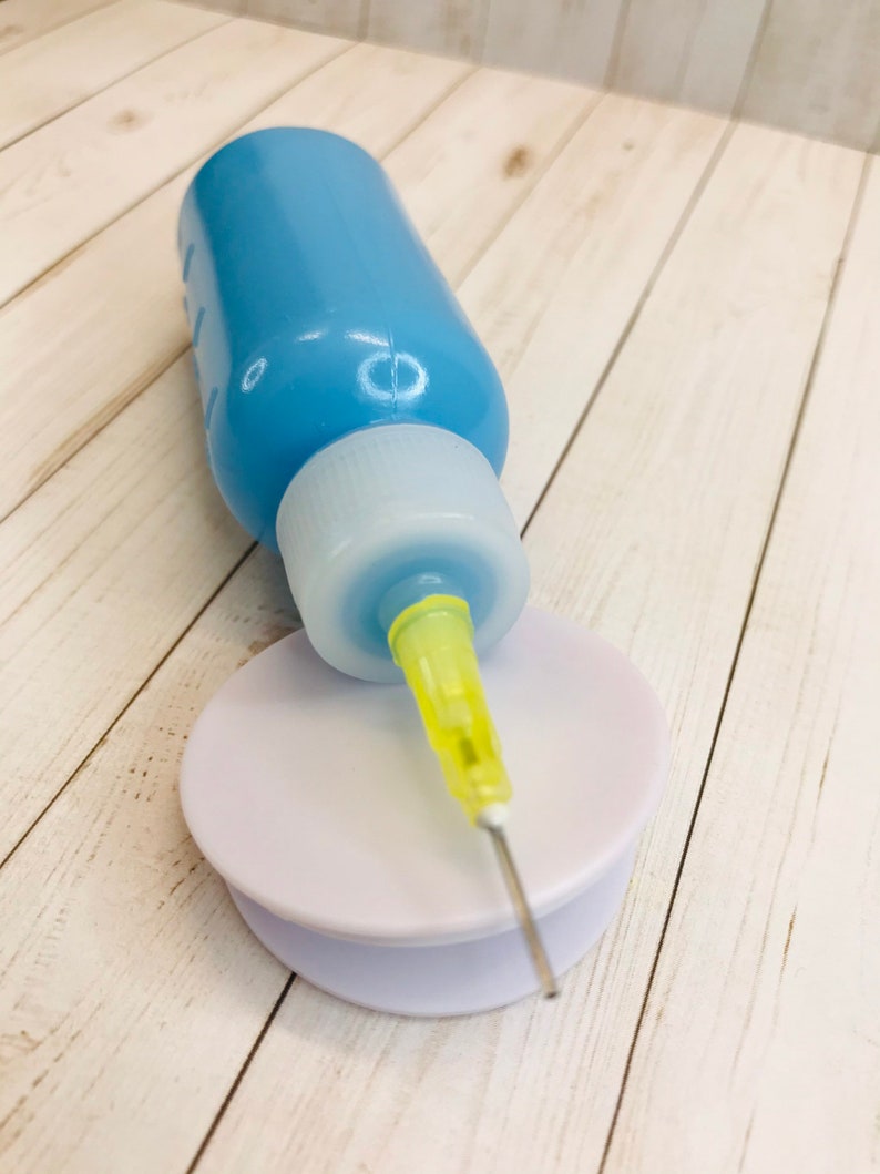 Op de afbeelding: Een blauwe plastic fles met een witte dop en een gele naald die aan de dop is bevestigd. De fles staat op een ronde witte basis.