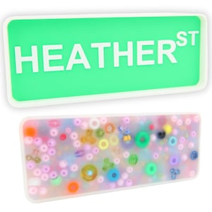 Pode incluir: Uma bandeja retangular verde com letras brancas que dizem "HEATHER ST". Uma segunda bandeja está cheia de contas e círculos coloridos.
