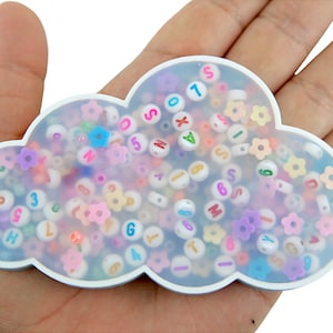 Könnte beinhalten: Ein weißer, wolkenförmiger Behälter gefüllt mit bunten Buchstaben- und Zahlenperlen. Die Perlen sind in verschiedenen Farben, darunter rosa, blau, gelb und lila.