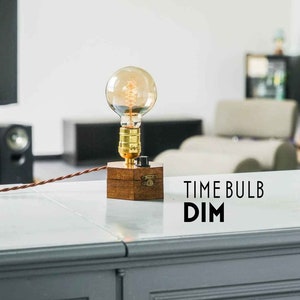 Könnte beinhalten: Eine Holzlampenbasis mit einer Messingfassung und einer klaren Glühbirne. Die Lampe ist an eine Steckdose angeschlossen und hat ein Kabel mit einem Schalter. Die Lampe steht auf einer grauen Arbeitsplatte mit dem Text "TIME BULB DIM" auf der Oberfläche.
