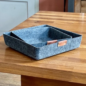 Könnte beinhalten: Zwei graue Filz-Aufbewahrungskörbe mit braunen Ledergriffen. Die Körbe sind ineinander verschachtelt und stehen auf einem Holztisch.