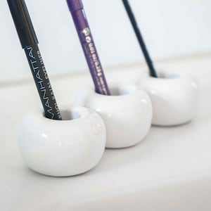Könnte beinhalten: Drei weiße Keramikhalter mit drei Make-up-Pinseln. Die Pinsel sind schwarz, lila und silber. Der schwarze Pinsel trägt den Text "MANHATTAN KHOL KAJAL EYELINER". Der lila Pinsel trägt den Text "88010 100 BLACK".
