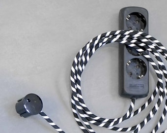 Op de afbeelding: Een zwart-wit gestreept stroomsnoer met een zwarte stekkerdoos met drie stopcontacten. De stekkerdoos heeft een zwarte kunststof behuizing en een wit label met zwarte tekst.