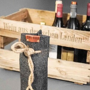 Könnte beinhalten: Ein dunkelgrauer Filz-Weinflaschenhalter mit einer Kordel aus Jute und einem kleinen orangefarbenen Etikett mit der Aufschrift "TIME BULB". Im Hintergrund eine Holzkiste mit Weinflaschen.