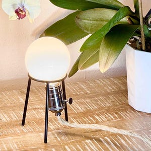 May include: A white globe lamp with a black metal tripod stand. The lamp is on a wooden surface and has a white cord.