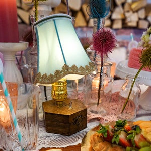 Pode incluir: Uma lâmpada de mesa de estilo vintage com um abajur azul claro e uma base de madeira. A lâmpada está sobre uma mesa com uma toalha de renda branca e um copo de água com um canudo listrado. Também há várias garrafas de vidro com flores e um prato de torta de frutas.