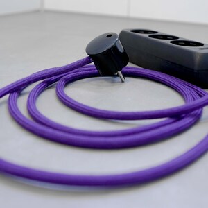 Może przedstawiać: Czarny rozgałęziacz z trzema gniazdami i fioletowym kablem pokrytym tkaniną. Kabel jest zwinięty, a rozgałęziacz jest podłączony.