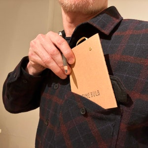 May include: A person wearing a black and red plaid shirt with a brown notebook in the pocket. The notebook has the text "TIME BULB" printed on the front.