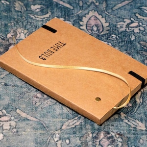 May include: A rectangular, kraft-colored notebook with the text "TIME BULB" printed on the cover. A gold ribbon is looped around the notebook. Two black elastic bands secure the notebook. A small gold rivet is visible.