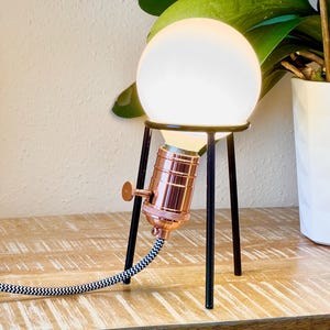 May include: A copper-colored table lamp with a white globe and a black tripod stand. The lamp has a black and white cord.