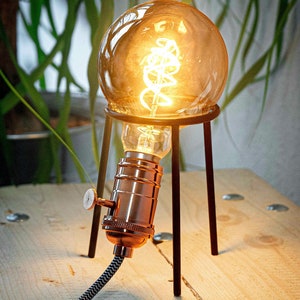 May include: A copper-colored light bulb with a coiled filament sits on a black metal stand with three legs. The light bulb is on and glowing. The stand is on a wooden surface.