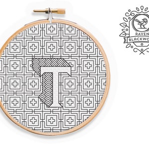 May include: A black and white embroidery hoop with a geometric pattern and the letter "T" in the center. The hoop is surrounded by a wooden frame. The text "RAVEN BLACKWORK" is visible in the upper right corner of the image.