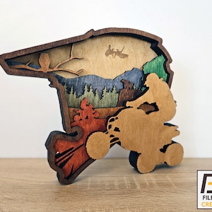 May include: A wooden cutout of a person riding an ATV through a forest scene. The ATV is brown and the forest scene is layered with different shades of blue, green, and brown. The cutout is mounted on a wooden base.