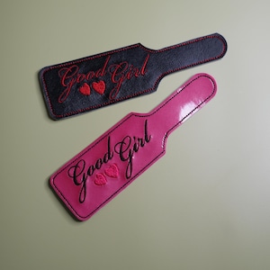 Pu&ograve; includere: Due pagaie, una nera e una rosa, ciascuna con il testo ricamato "Good Girl" in corsivo. La pagaia nera ha cuciture e cuori rossi, mentre la pagaia rosa ha cuciture e cuori neri. Le pagaie sono su uno sfondo verde chiaro.