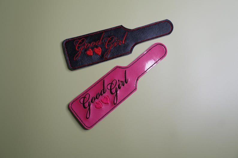 Pu&ograve; includere: Due pagaie, una nera e una rosa, ciascuna con il testo ricamato "Good Girl" in corsivo. La pagaia nera ha cuciture e cuori rossi, mentre la pagaia rosa ha cuciture e cuori neri. Le pagaie sono su uno sfondo verde chiaro.