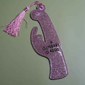 May include: A pink glitter bookmark with a tassel. The bookmark is shaped like a hand with the text "A Cliterary Classic" in black. The tassel is pink and attached to the top of the bookmark.